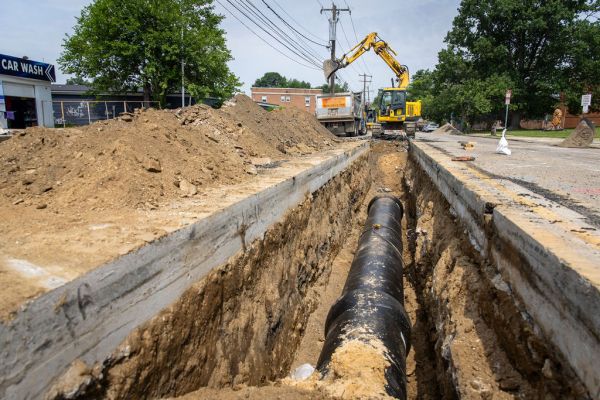 New water transmission main being installed. The camera looks down a road with one lane excavated, the double yellow line visible just to the right, dirt mounded along the left side, the excavator sits at the end of the trench extending it, and a new section of main sitting in the trench.