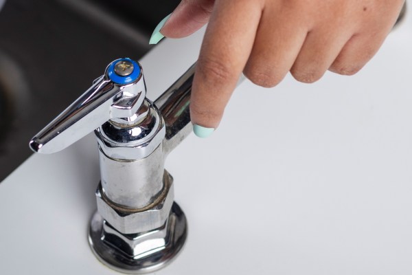Close-up of a hand with warm medium-brown skin and long, mint-colored fingernails, reaching for the handle of a sink faucet.
