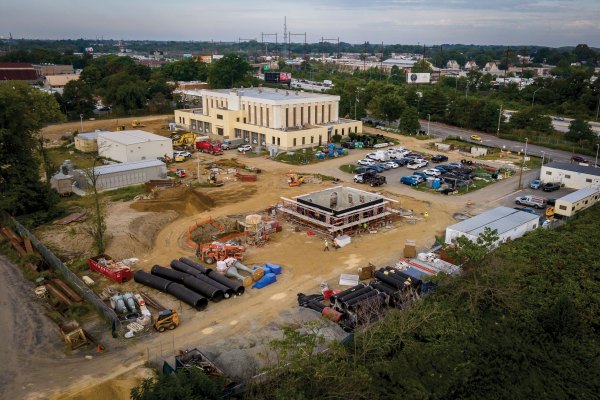 Aerial view of the Torresdale Pump Station under construction, with heavy machinery, orange temporary fencing, and segments of large black piping stacked on the perimeter.