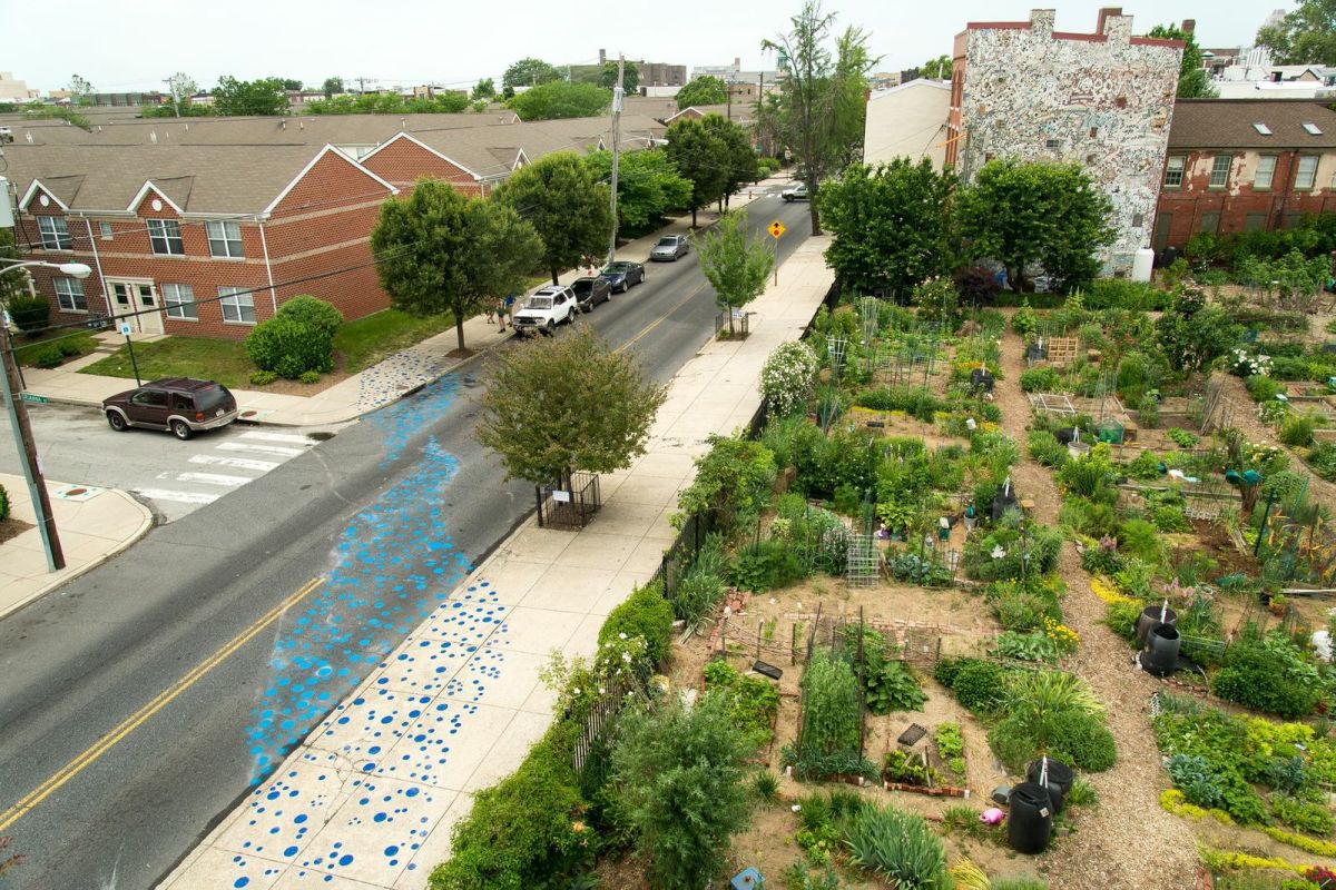 elevated view of a Philadelphia block with a community garden divided into neat square plots on the right, stormwater trees between the street and sidewalk, and a water art installation featuring a constellation of blue dots painted on the street and sidewalk in an undulating diagonal line mimicking a stream and housing visible in the upper left corner.