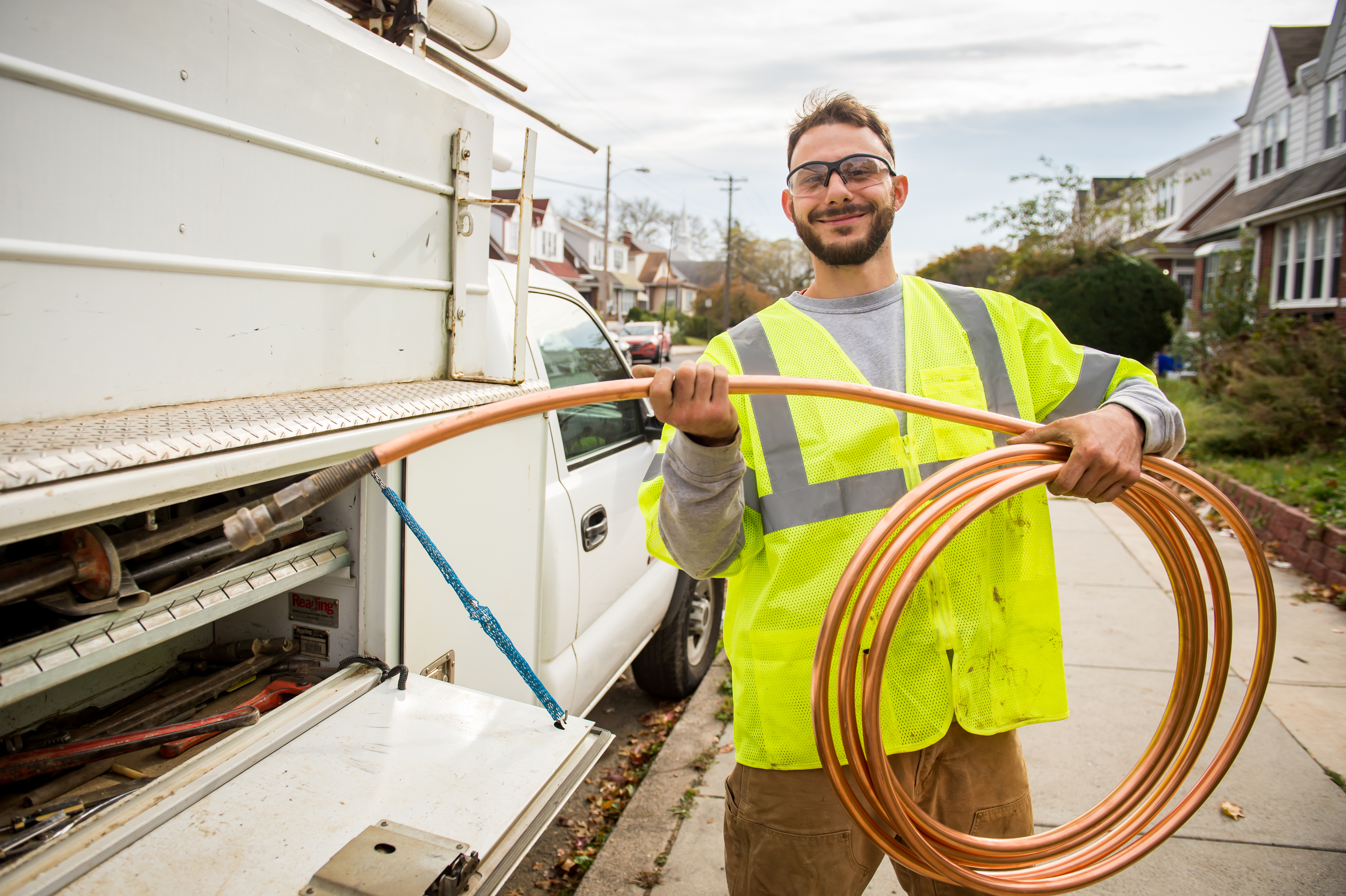 A worker with short, light brown hair and facial hair, wearing safety glasses and a neon yellow safety vest, stands smiling in front of a white work truck holding up a coil of new copper piping.