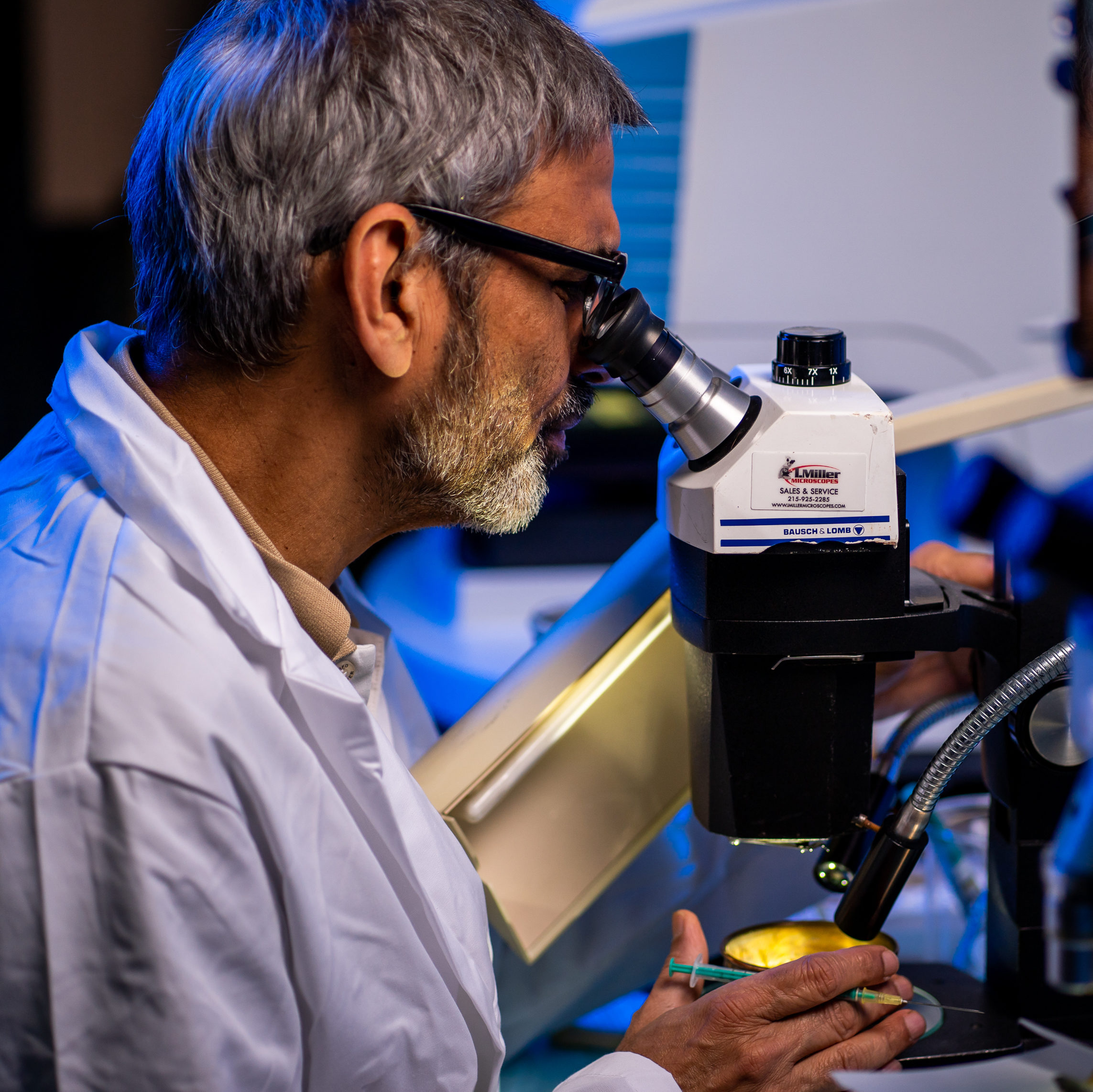 a scientist with short salt and pepper hair and close cropped beard, wearing a white lab coat and black glasses is seen in profile, peering into a microscope with pencil in hand.