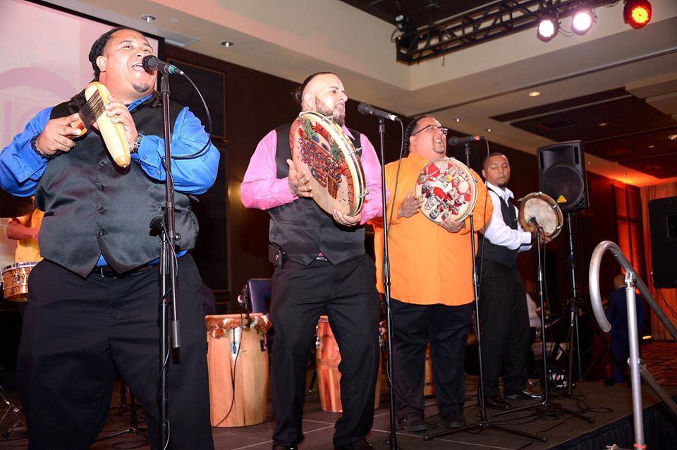 Los Bomberos De la Calle on stage, with lights shining down. Each member holds some sort of hand percussion, with the three closest to the camera standing angled towards us, singing into microphones. Speakers and more drums are visible behind them.