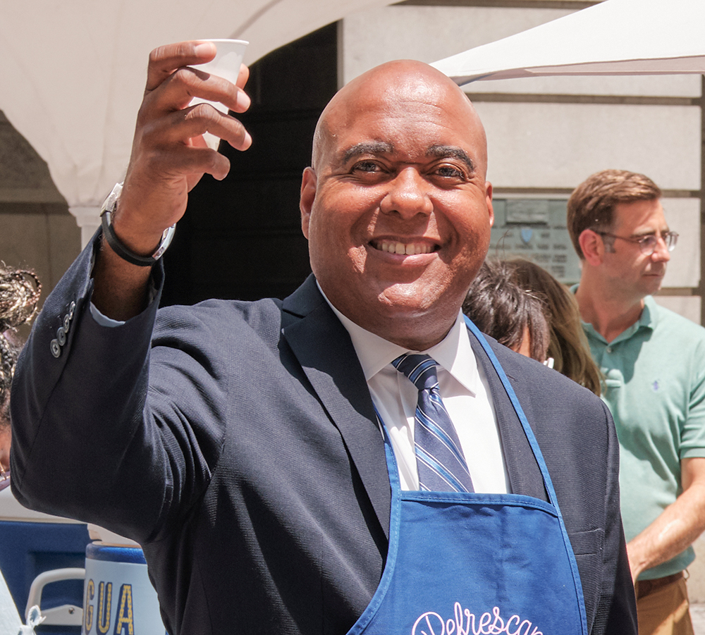 PWD Commissioner Randy Hayman stands smiling in the courtyard at city hall, wearing a blue apron, as he raises a paper cone of fresh, cold, Philly tap water to toast Philly tap after serving customers and passers-by at Philly Water Bar.