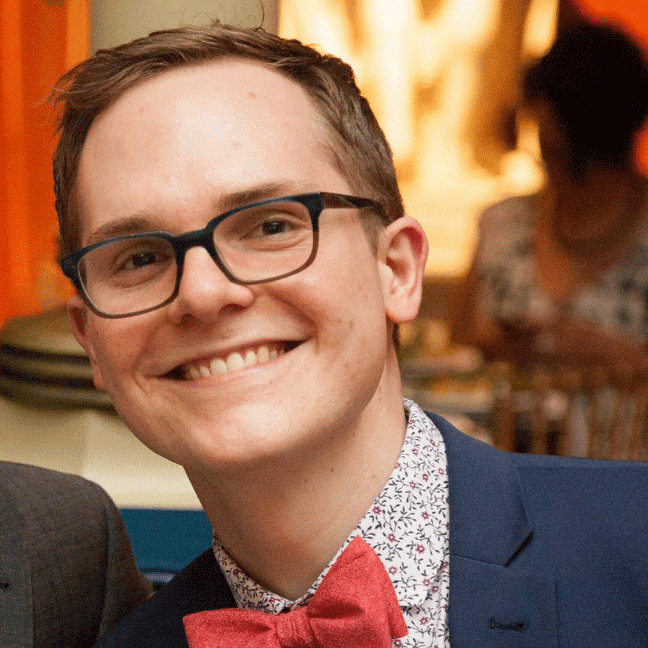 Herbie is photographed at an event wearing a navy blue suit with white patterned shirt and a snazzy red bow tie, with black rectangular glasses and a cheerful smile.