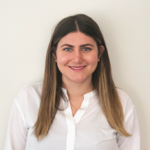 Hailey's headshot shows her standing smiling in front of a cream colored wall wearing a white button-down shirt, with her long brown hair draped in front of her shoulders.