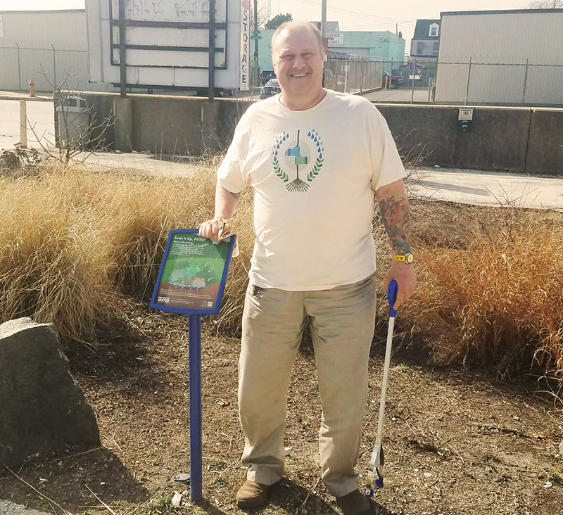 John is a smiling, tattooed gentlman seen leaning on a Soak It Up Adoption info sign in one of the Eadom Street rain gardens, wearing a t-shirt that features the Adoption Program 'crest', two different garden-gloved hands coming together to grasp a rake, encircled by leaves and raindrops, and holding a trash grabber